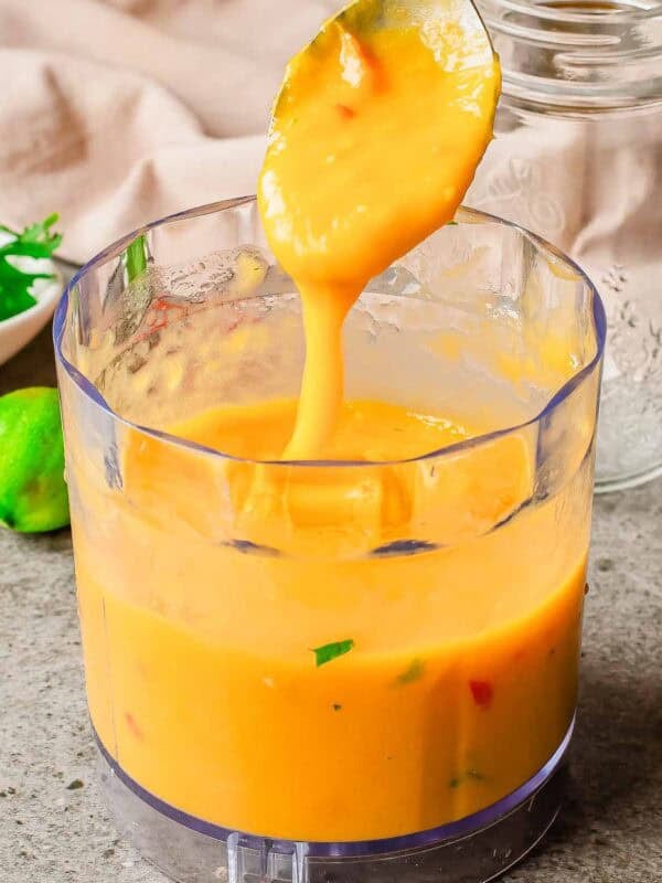 A spoon lifts thick, creamy mango sauce from a clear food processor bowl. Some green and red flecks are visible in the sauce. The background shows a lime, herbs, and a glass jar on a gray countertop.
