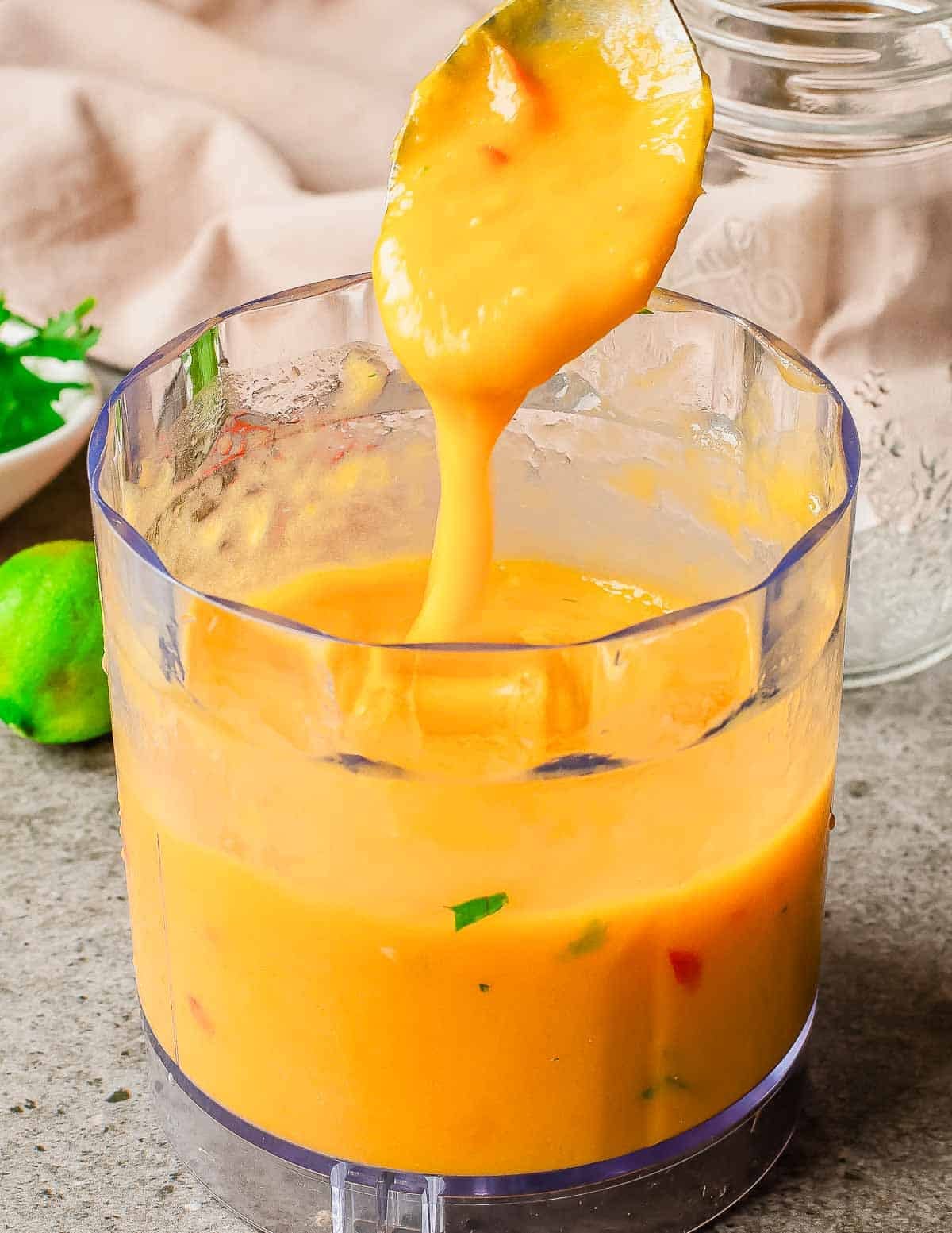 A spoon lifts thick, creamy mango sauce from a clear food processor bowl. Some green and red flecks are visible in the sauce. The background shows a lime, herbs, and a glass jar on a gray countertop.