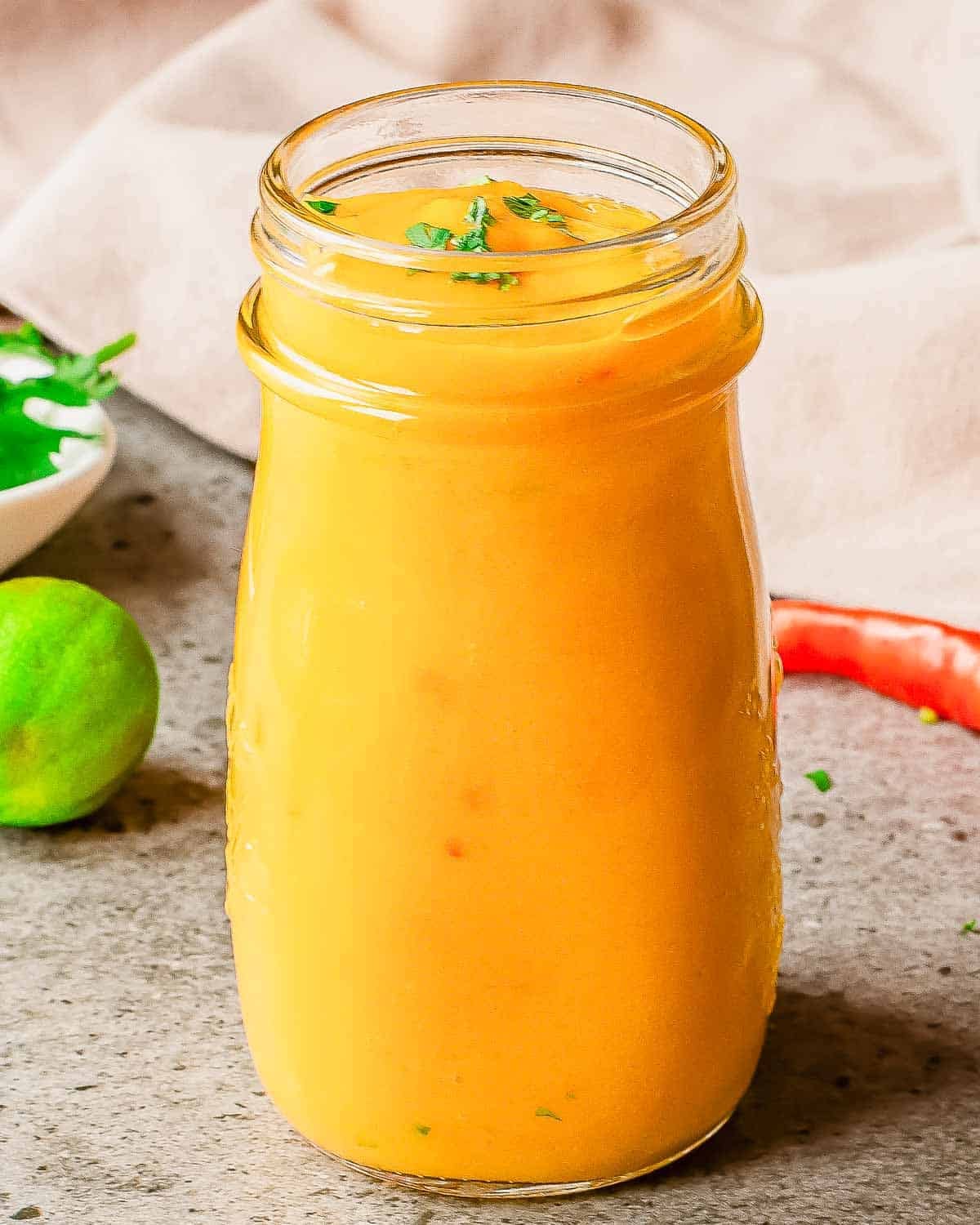 A glass jar filled with mango salad sauce, garnished with chopped herbs. The jar is on a gray surface with a lime and a red pepper in the background.