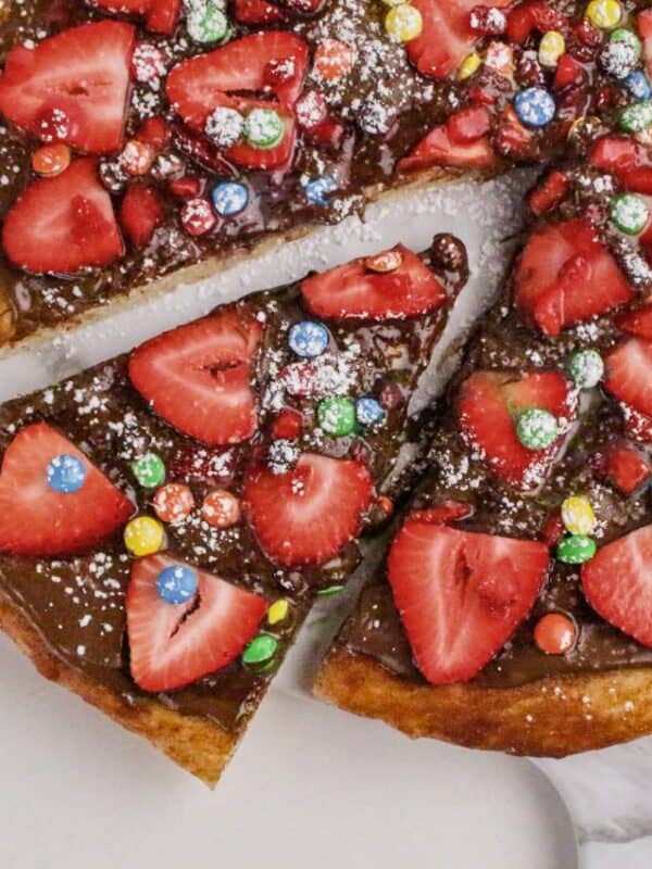 A dessert pizza topped with sliced strawberries, colorful candy-coated chocolates, and powdered sugar on a rich chocolate spread—one slice being lifted—makes this a standout among irresistible Nutella dessert recipes.