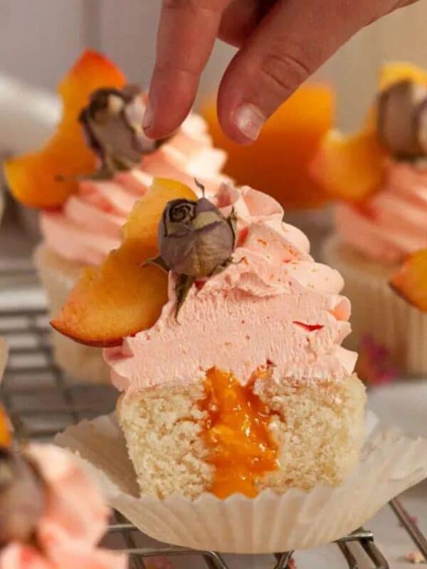 A hand decorates peach cupcakes topped with pink frosting, peach slices, and dried rosebuds. One cupcake is cut open, revealing a peach filling inside—an irresistible upgrade for your dessert table and inspiration for new cupcake recipes.