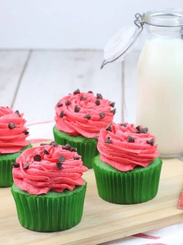 Four cupcakes with pink frosting and chocolate chips in green wrappers are on a wooden board next to a glass bottle of milk and red polka dot straws, on a white surface with a striped towel.