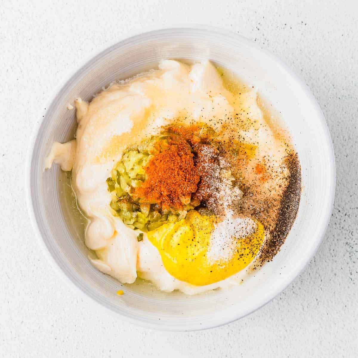 A white bowl containing mayonnaise, chopped pickles, yellow mustard, paprika, black pepper, salt, and other seasonings on a white surface.