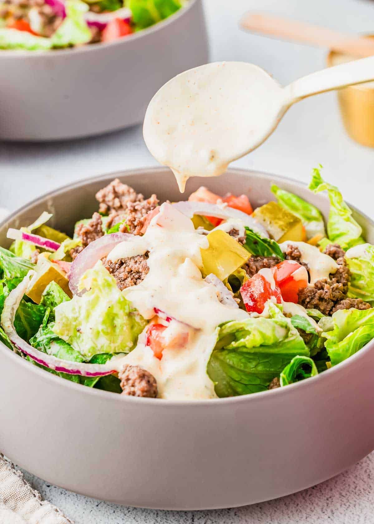 A gray bowl filled with a salad containing ground beef, lettuce, tomato, pickles, and red onions, with creamy dressing being drizzled on top from a spoon above.