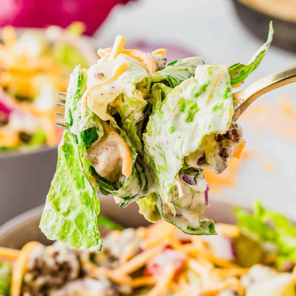 A close-up of a fork holding a bite of salad with leafy greens, ground beef, shredded cheddar cheese, and creamy dressing. The background shows more salad in a bowl.