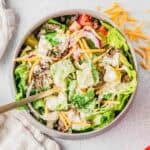 A bowl of salad filled with lettuce, ground beef, shredded cheddar cheese, sliced red onions, diced tomatoes, pickles, and creamy dressing, with a fork in the bowl and some cheese scattered on the table.