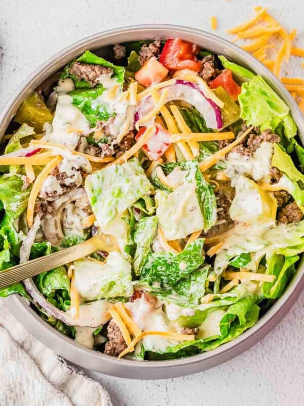 A bowl of salad filled with lettuce, ground beef, shredded cheddar cheese, sliced red onions, diced tomatoes, pickles, and creamy dressing, with a fork in the bowl and some cheese scattered on the table.