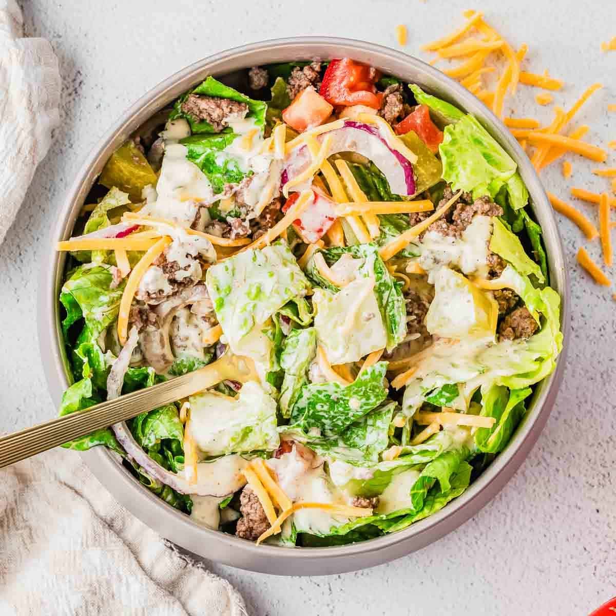 A bowl of salad filled with lettuce, ground beef, shredded cheddar cheese, sliced red onions, diced tomatoes, pickles, and creamy dressing, with a fork in the bowl and some cheese scattered on the table.