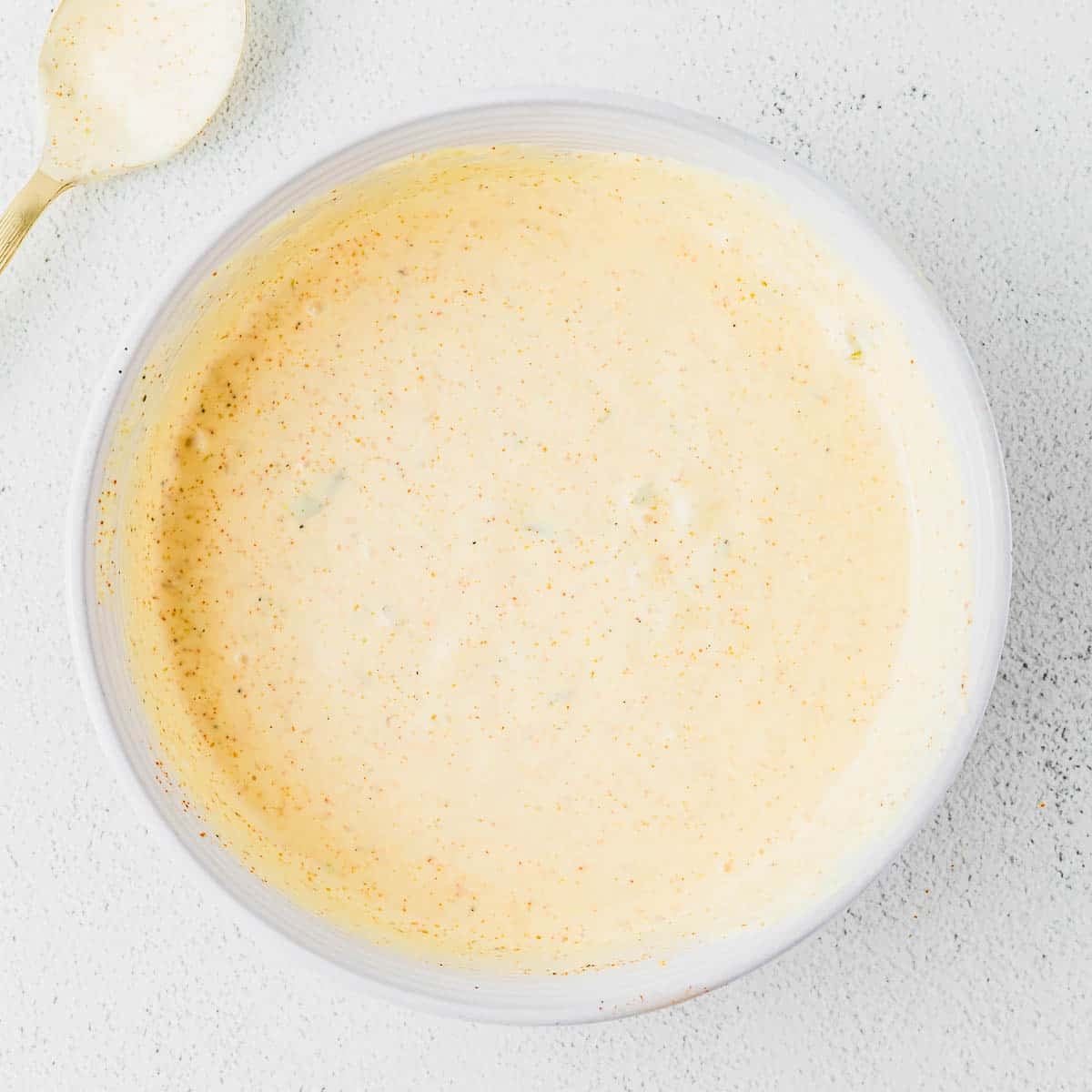 A white bowl filled with a creamy, light burger sauce with specks of seasoning, placed on a light textured surface. A gold spoon with some sauce remains visible in the upper left corner.