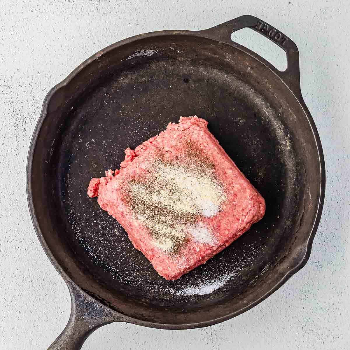 A square portion of raw ground beef with salt and pepper sprinkled on top sits in a black cast iron skillet on a light gray surface.