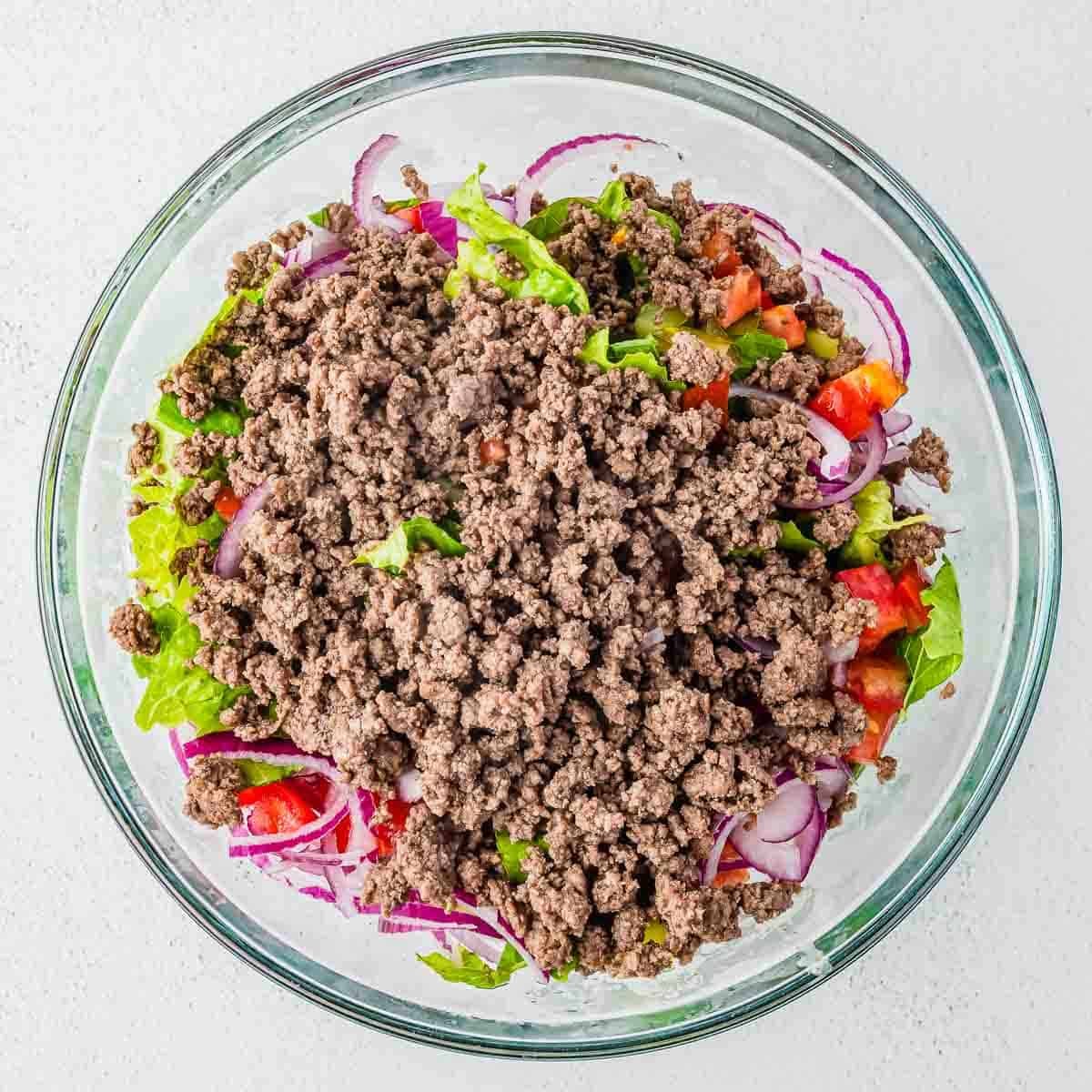 A glass bowl filled with cooked ground beef on top of a salad with lettuce, sliced red onions, and diced tomatoes, all placed on a light-colored surface.