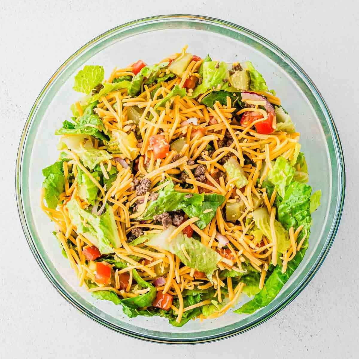 A glass bowl filled with a colorful Big Mac burger salad containing lettuce, ground beef, shredded cheese, diced tomatoes, and tortilla chips.