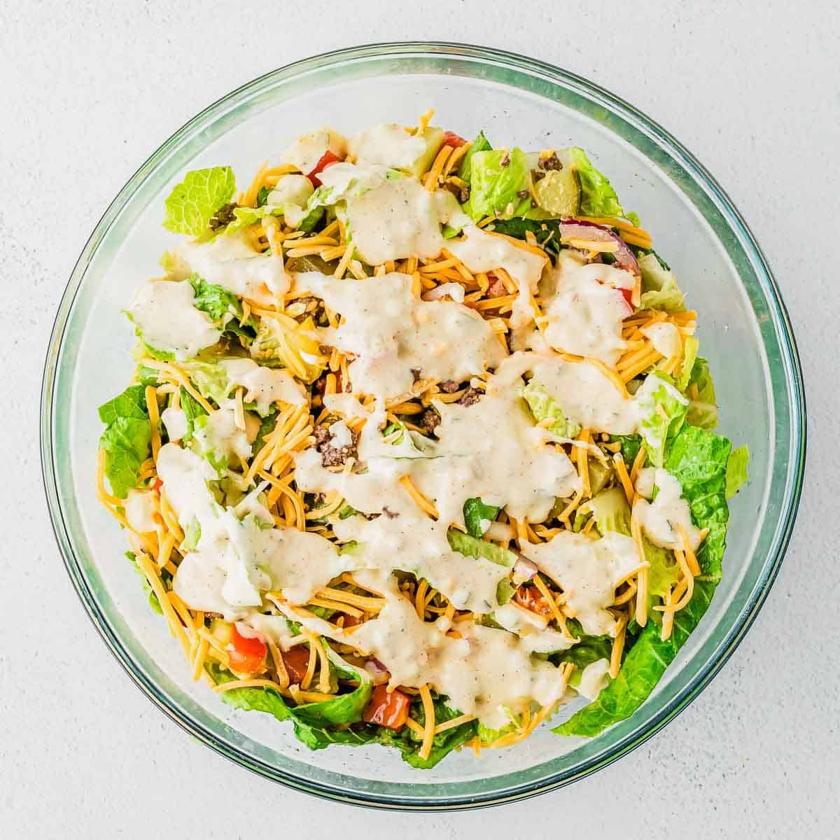 A clear glass bowl filled with chopped lettuce, shredded cheddar cheese, tomatoes, and creamy dressing, on a white surface.