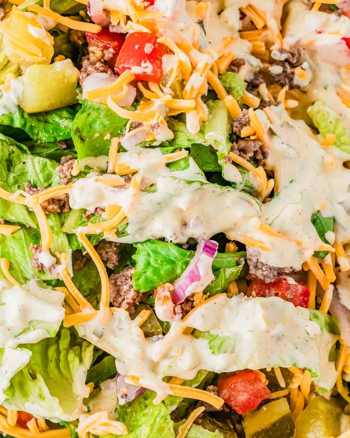 A close-up of a fresh Big Mac salad with chopped lettuce, diced tomatoes, shredded cheddar cheese, ground beef, red onions, pickles, and a creamy dressing drizzled on top.