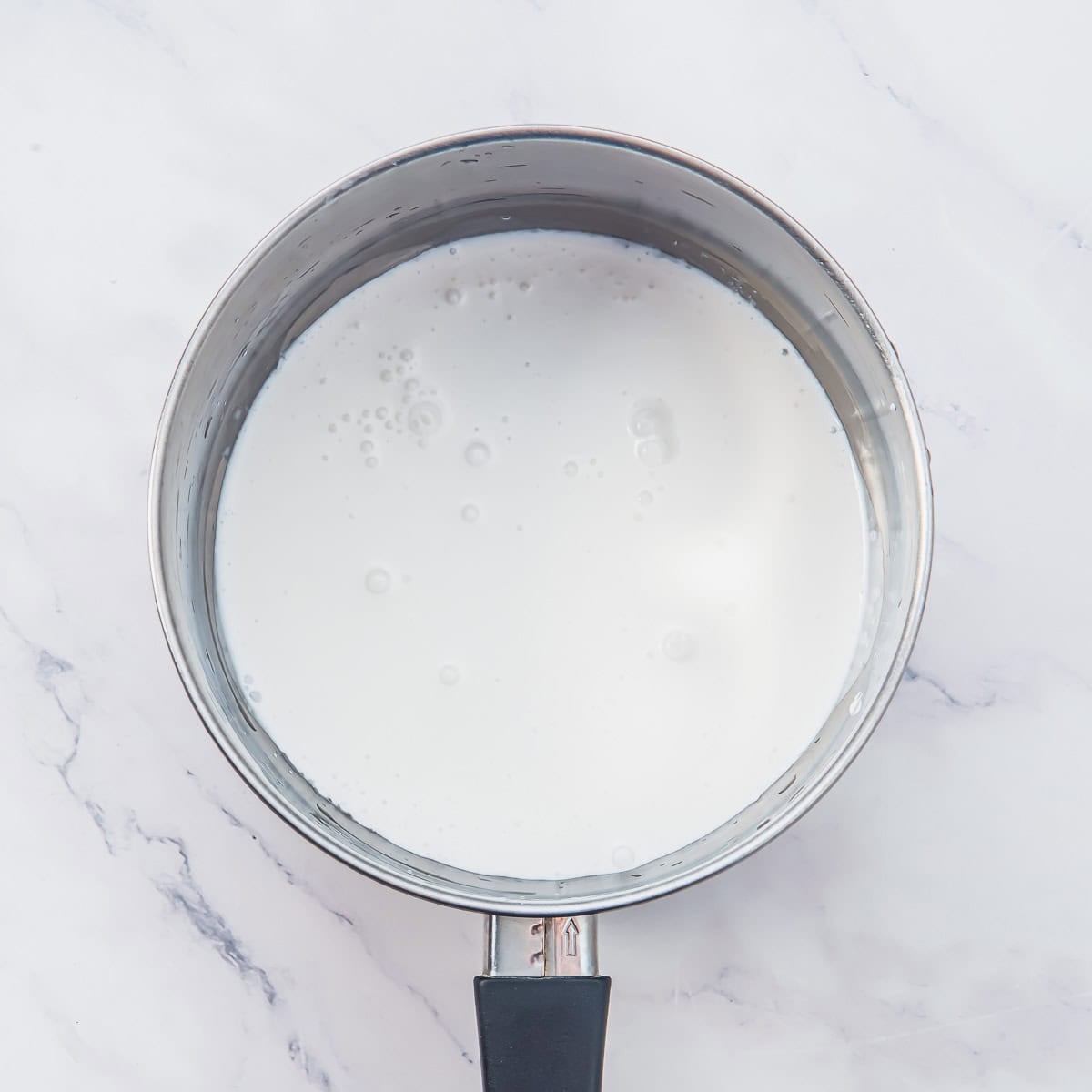A stainless steel saucepan with a black handle, filled with coconut milk sits on a white marble surface.