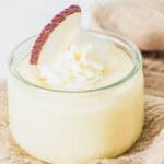 A small glass jar filled with creamy yellow coconut pudding, topped with a swirl of whipped cream and a slice of fresh coconut, placed on a beige textured cloth.