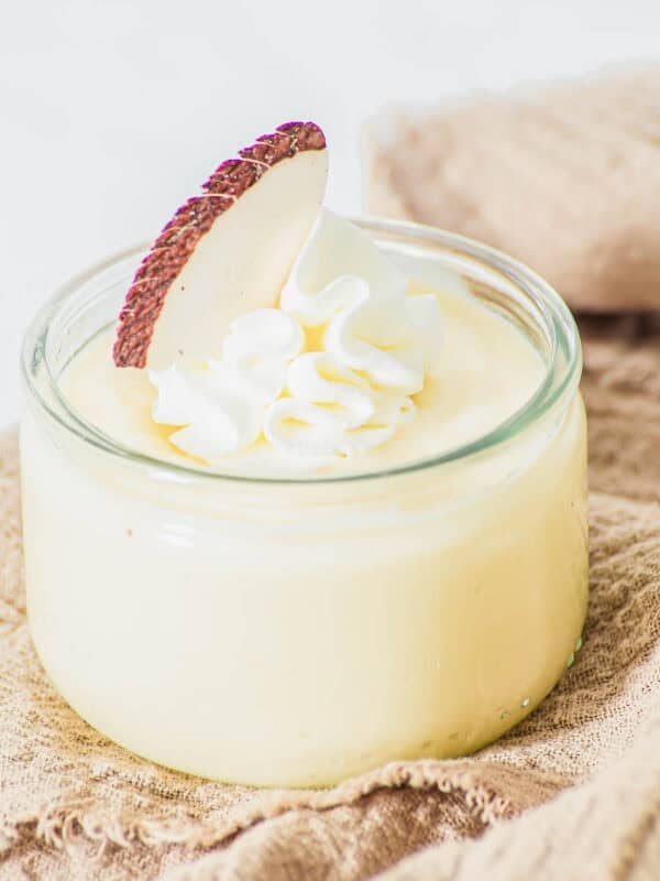 A small glass jar filled with creamy yellow coconut pudding, topped with a swirl of whipped cream and a slice of fresh coconut, placed on a beige textured cloth.