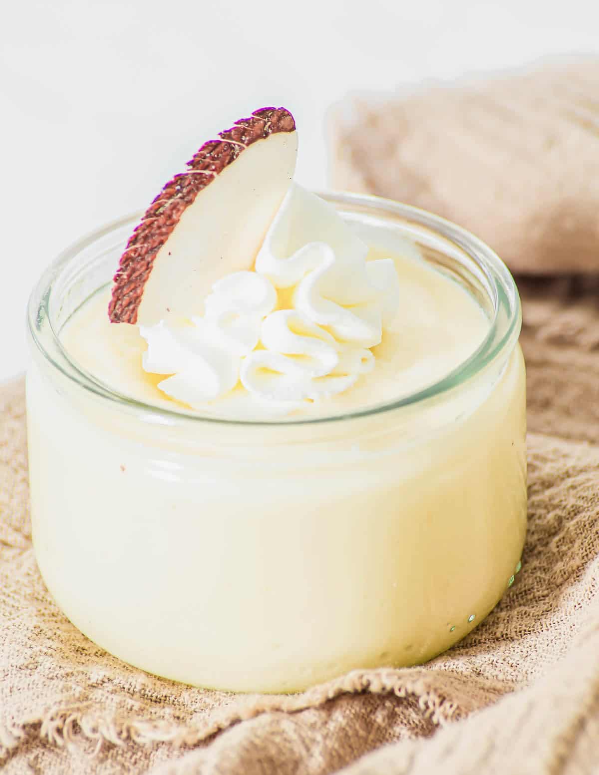 A small glass jar filled with creamy yellow coconut pudding, topped with a swirl of whipped cream and a slice of fresh coconut, placed on a beige textured cloth.