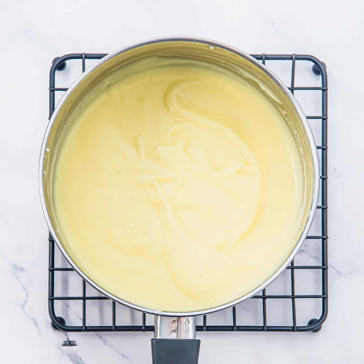 A saucepan filled with smooth, pale yellow pudding sits on a black wire cooling rack over a white marble surface.