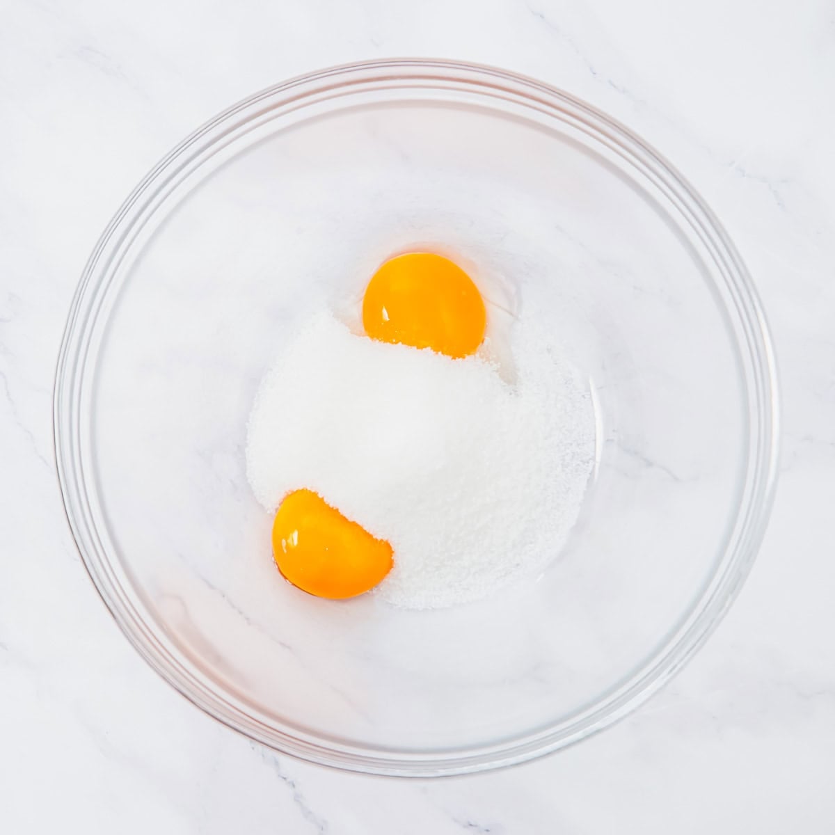 A glass bowl containing two egg yolks and a mound of granulated sugar, placed on a white marble surface.