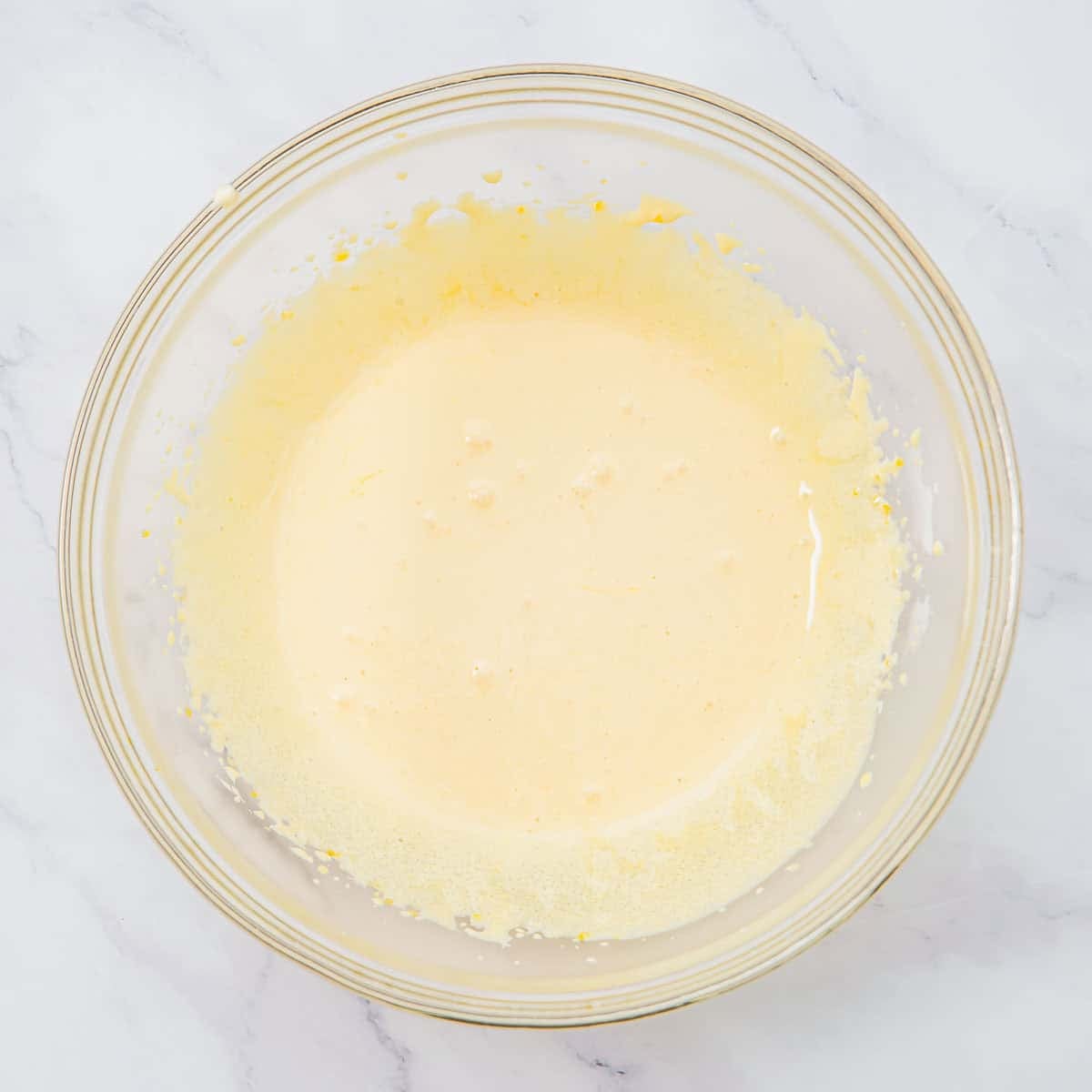 A glass bowl filled with whisked sugar and egg yolk mixture sits on a white marble surface. Small bubbles and flecks are visible around the edges of the mixture.