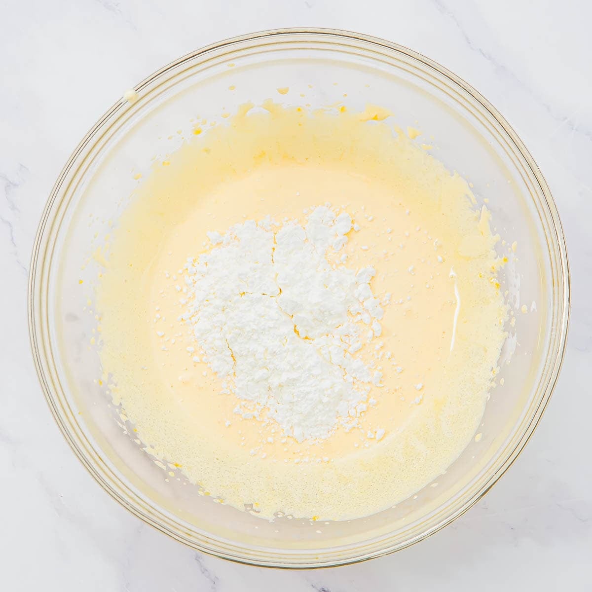 A glass bowl with whisked egg yolks and sugar with a pile of cornstarch in the center, on a white marble surface.