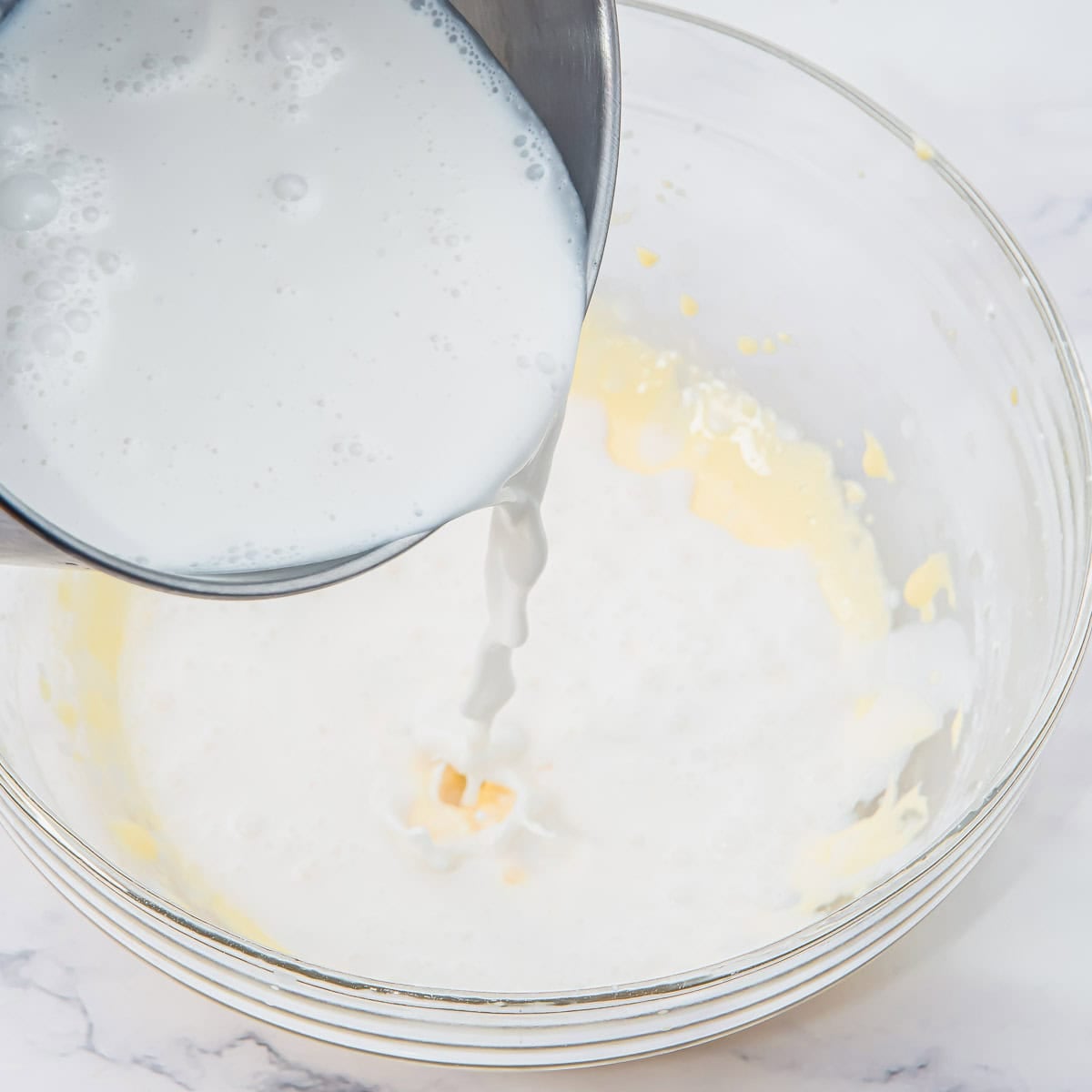 A metal bowl pours coconut milk into a glass mixing bowl containing a light yellow mixture, on a white marble surface.