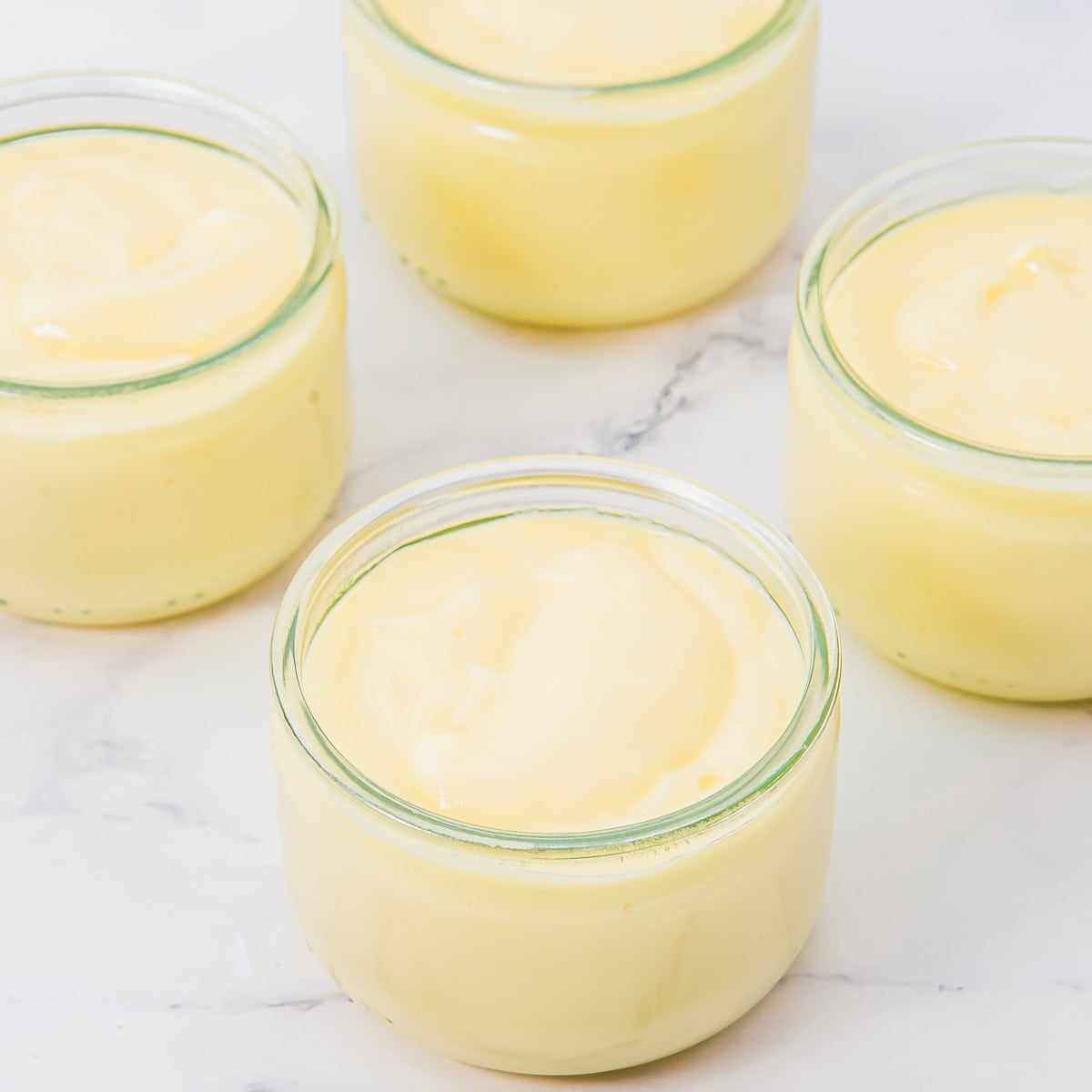Four glass jars filled with creamy yellow coconut pudding are arranged on a white marble surface. The pudding has a smooth, thick texture and each jar is filled nearly to the top.