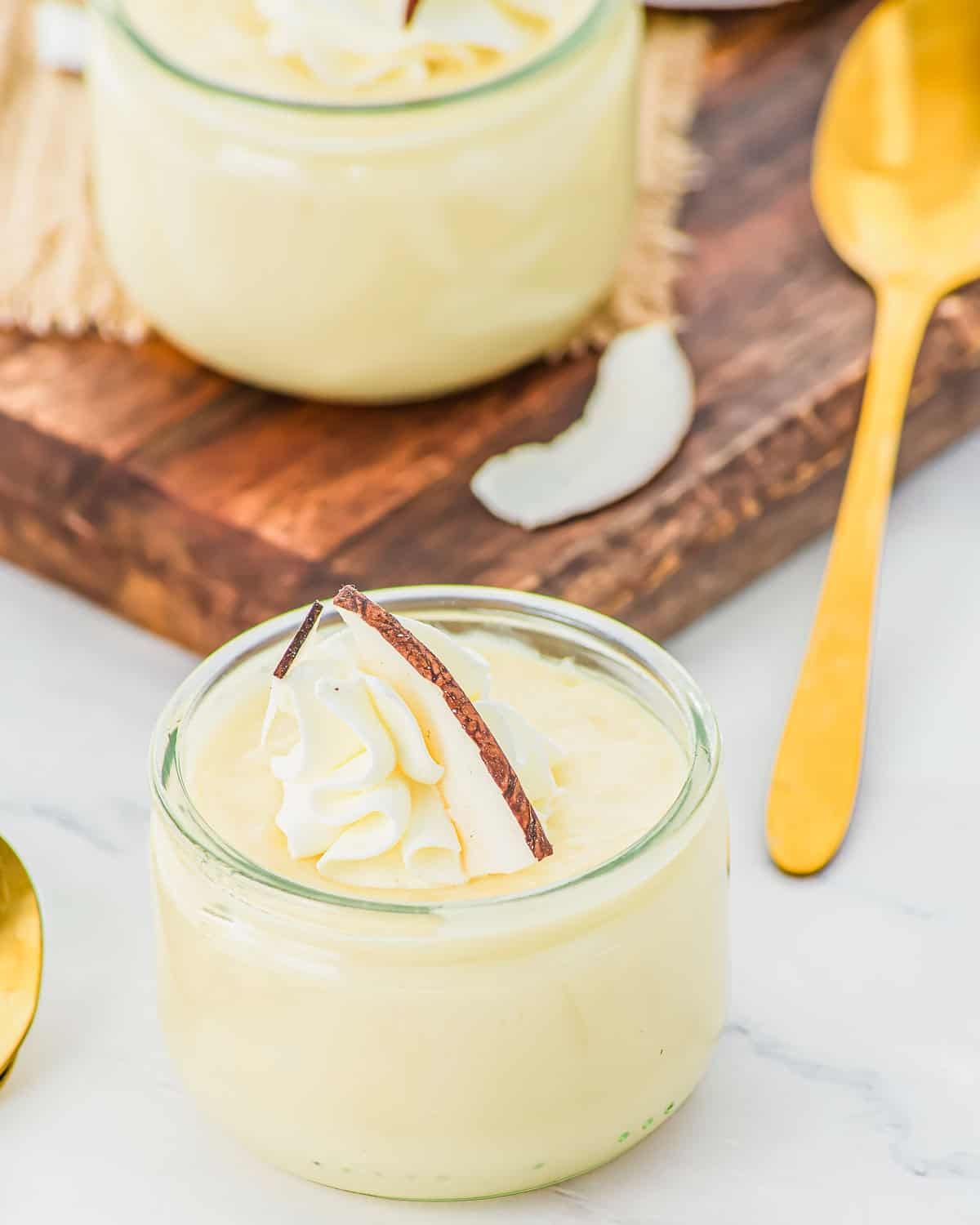 A small glass jar filled with creamy coconut pudding, topped with whipped cream and a slice of coconut, sits next to a gold spoon on a light surface. Another jar and a wooden board are in the background.