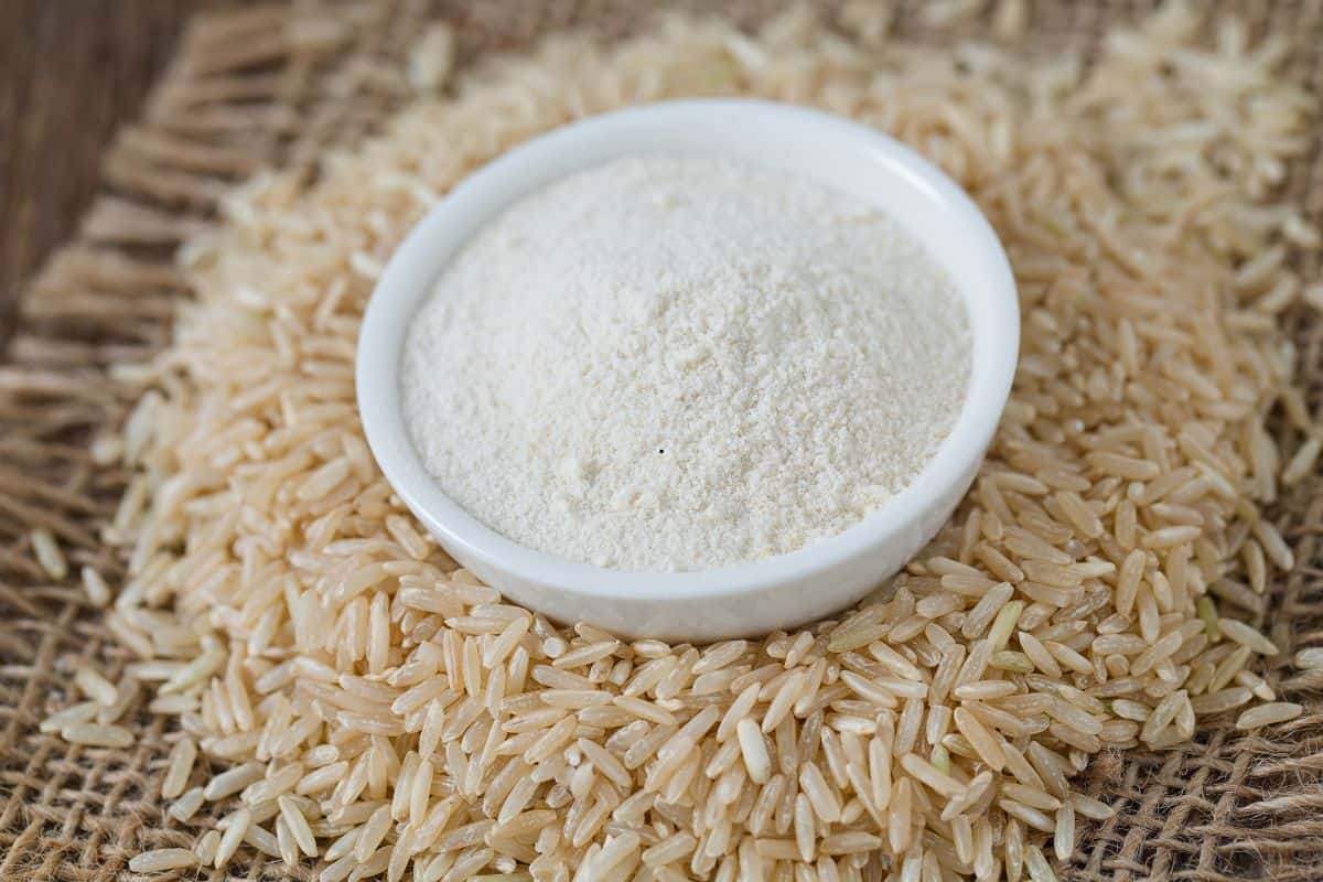 A small white bowl filled with rice flour, a popular substitute for cornstarch, sits on a pile of uncooked brown rice grains, all placed on a piece of burlap fabric.