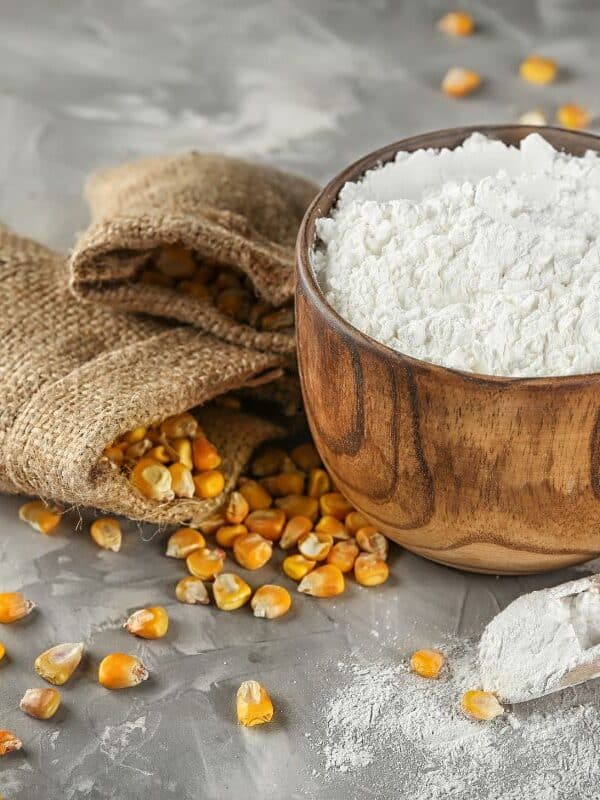 A wooden bowl filled with corn flour, a popular substitute for cornstarch, sits on a gray surface next to a small wooden scoop of flour, a burlap sack, and scattered yellow corn kernels.