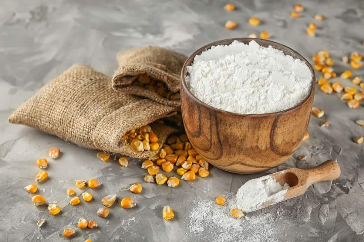 A wooden bowl filled with corn flour, a popular substitute for cornstarch, sits on a gray surface next to a small wooden scoop of flour, a burlap sack, and scattered yellow corn kernels.