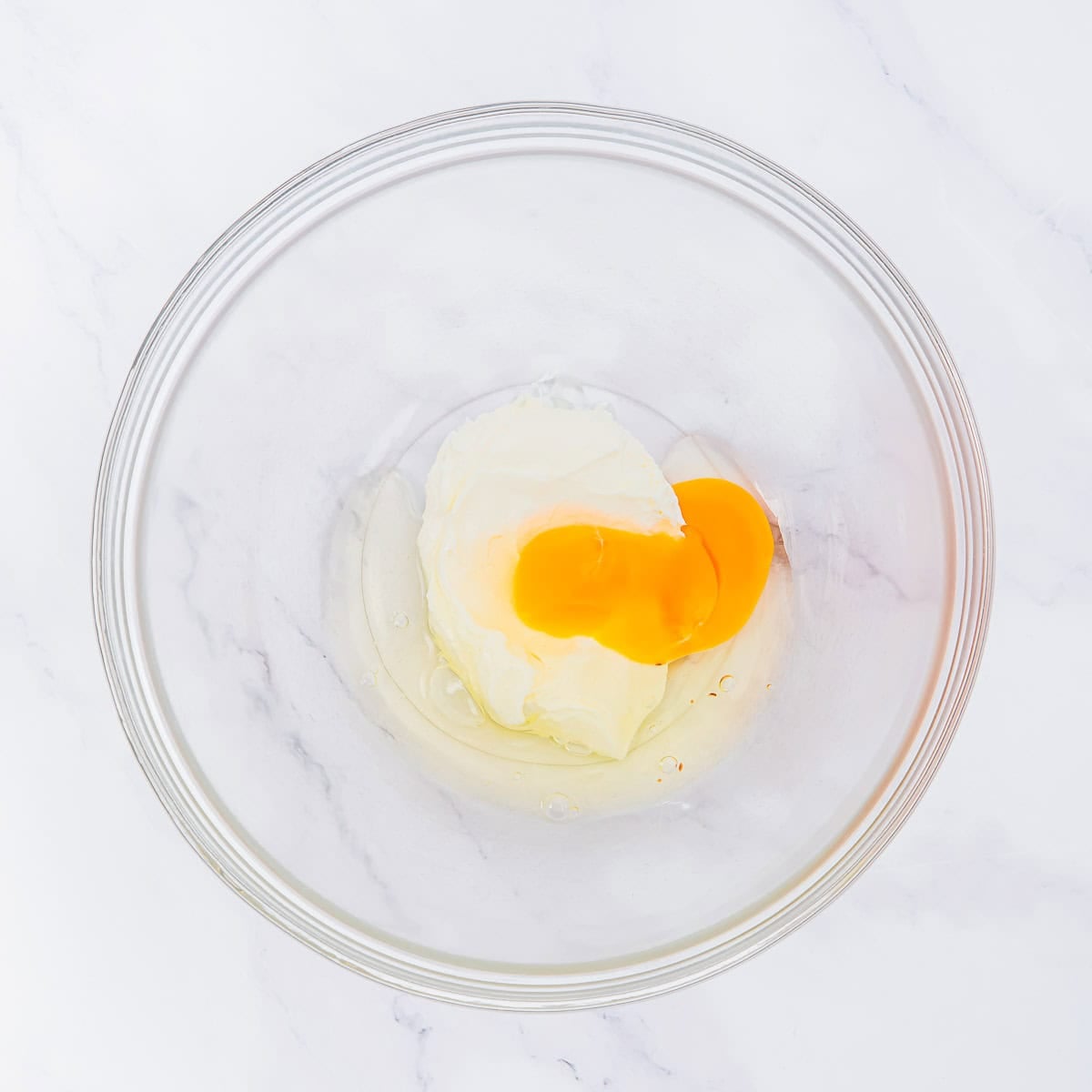 A glass mixing bowl containing cream cheese and raw eggs on a white marble surface.