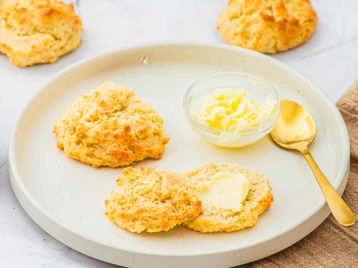 A round plate with two golden-brown almond flour biscuits, one split and spread with butter, next to a small glass bowl of butter and a gold spoon. More biscuits are visible in the background.