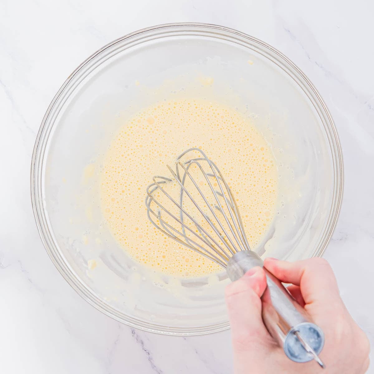 A hand is holding a whisk and mixing a pale yellow batter in a clear glass bowl on a white marble surface.