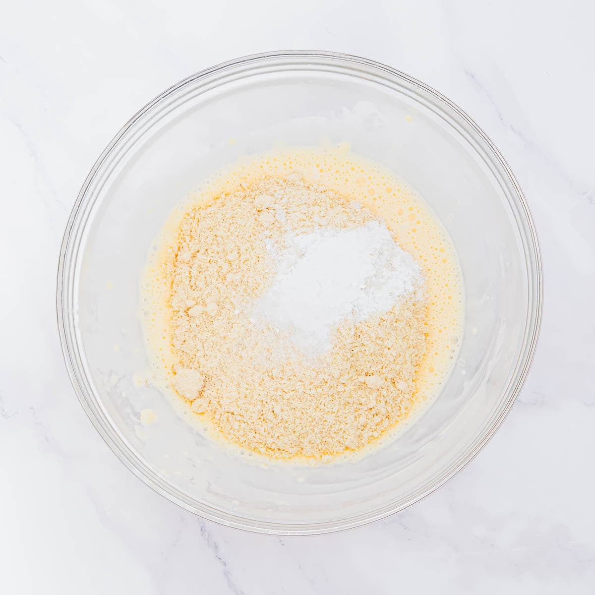 A clear glass bowl containing a yellow, beaten mixture with dry almond flour and a mound of white baking powder on top, set on a white marble surface.