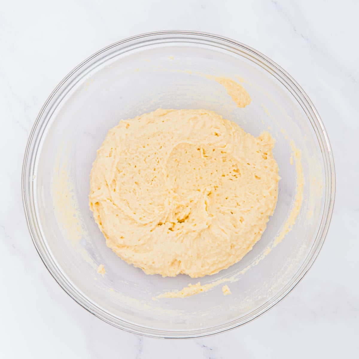 A glass mixing bowl filled with thick, creamy biscuit batter sits on a white marble surface.