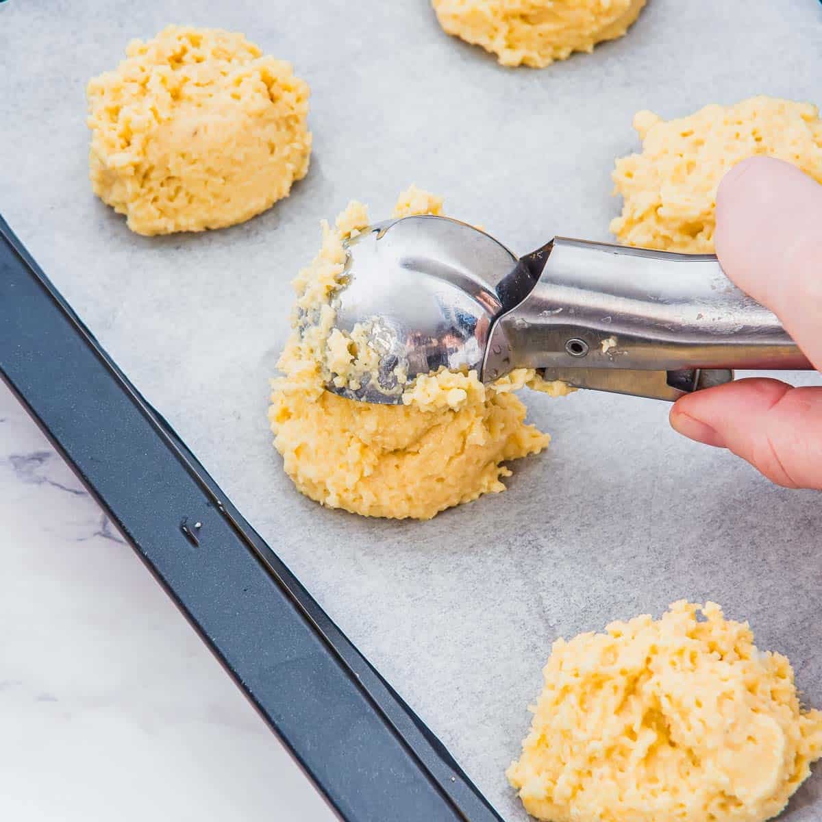 A hand uses an ice cream scoop to place balls of biscuit dough onto a parchment-lined baking tray; several dough portions are already on the tray, ready for baking.