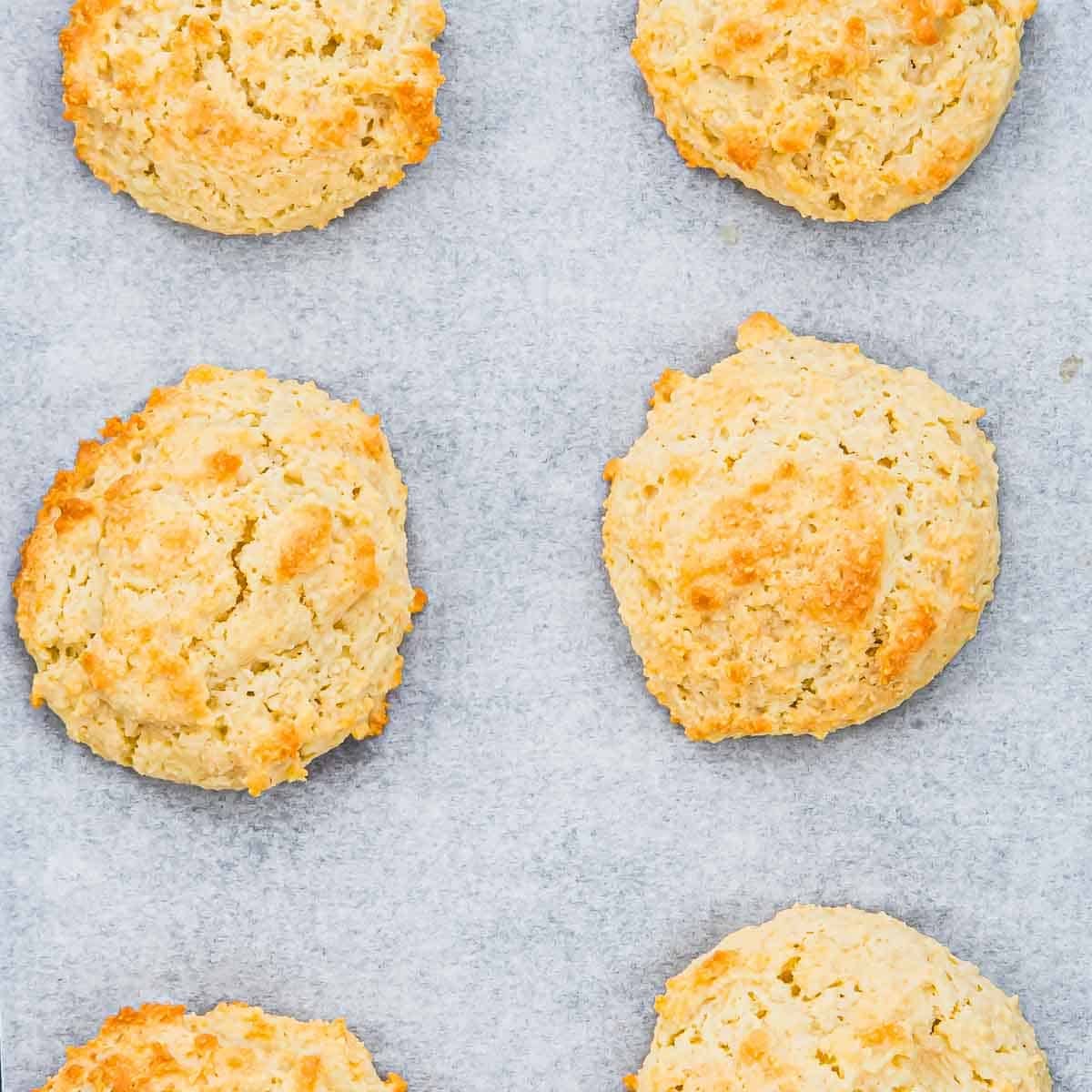 Six golden-brown almond flour drop biscuits are arranged on a sheet of parchment paper, spaced evenly apart, showing their rough, textured tops and slightly crisp edges.