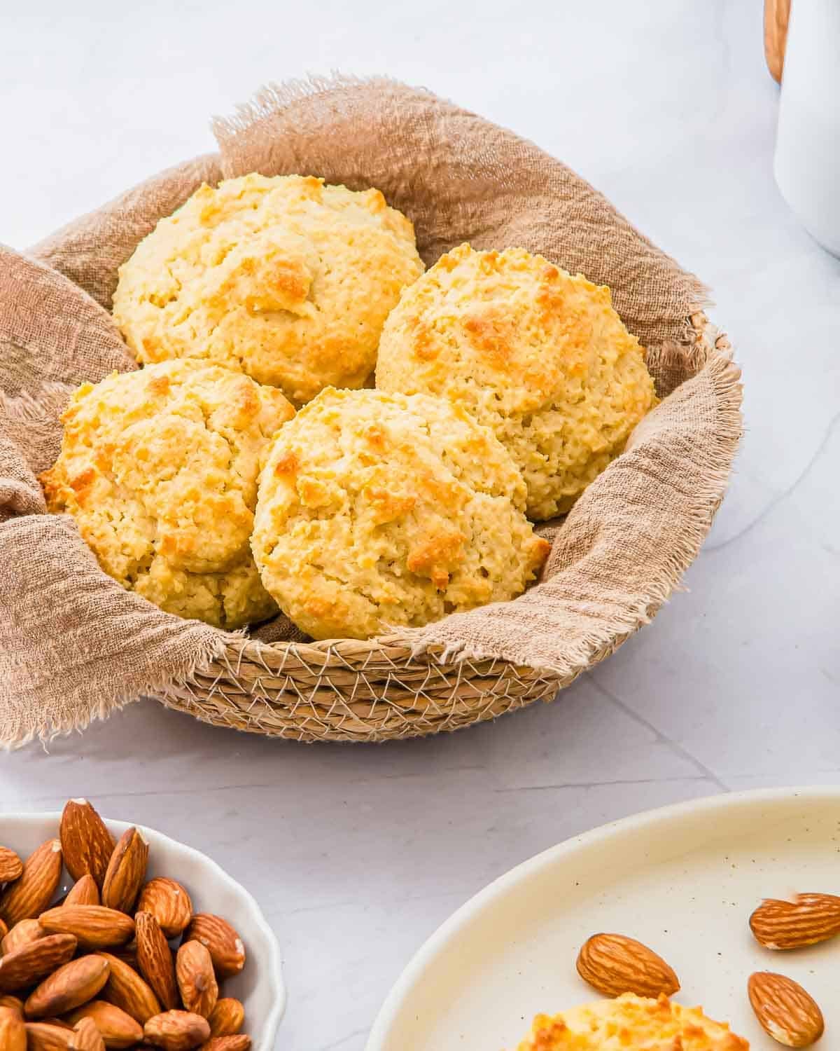 A basket lined with brown cloth holds four golden, freshly baked almond flour biscuits. Nearby, a small plate of whole almonds and part of a white plate are visible on a light marble surface.