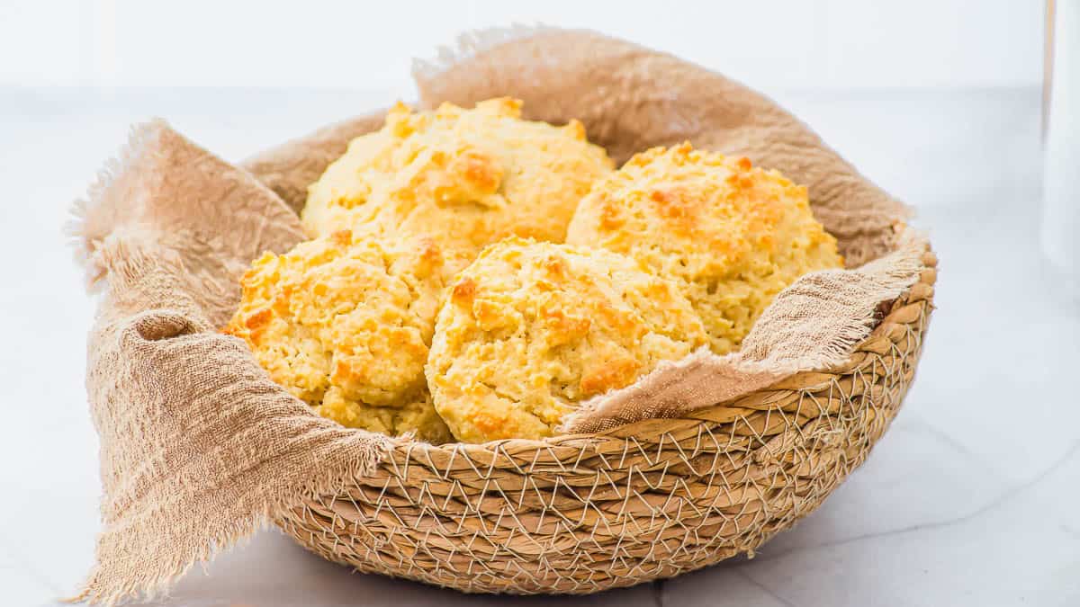 A woven basket lined with a beige cloth holds several golden-brown, freshly baked almond flour biscuits on a light surface.