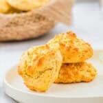 Two golden-brown drop biscuits sit stacked on a white plate, with a basket of more biscuits in the background. The biscuits have a crisp, textured surface and a light, fluffy appearance.