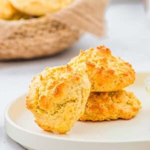Two golden-brown drop biscuits sit stacked on a white plate, with a basket of more biscuits in the background. The biscuits have a crisp, textured surface and a light, fluffy appearance.