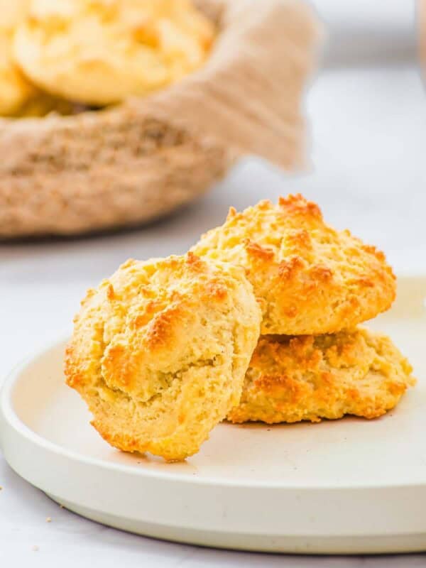Two golden-brown drop biscuits sit stacked on a white plate, with a basket of more biscuits in the background. The biscuits have a crisp, textured surface and a light, fluffy appearance.