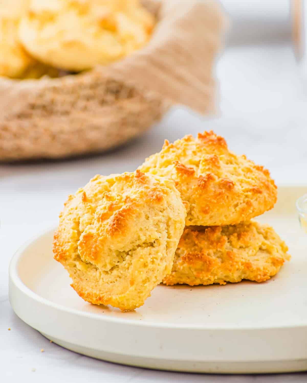Two golden-brown drop biscuits sit stacked on a white plate, with a basket of more biscuits in the background. The biscuits have a crisp, textured surface and a light, fluffy appearance.