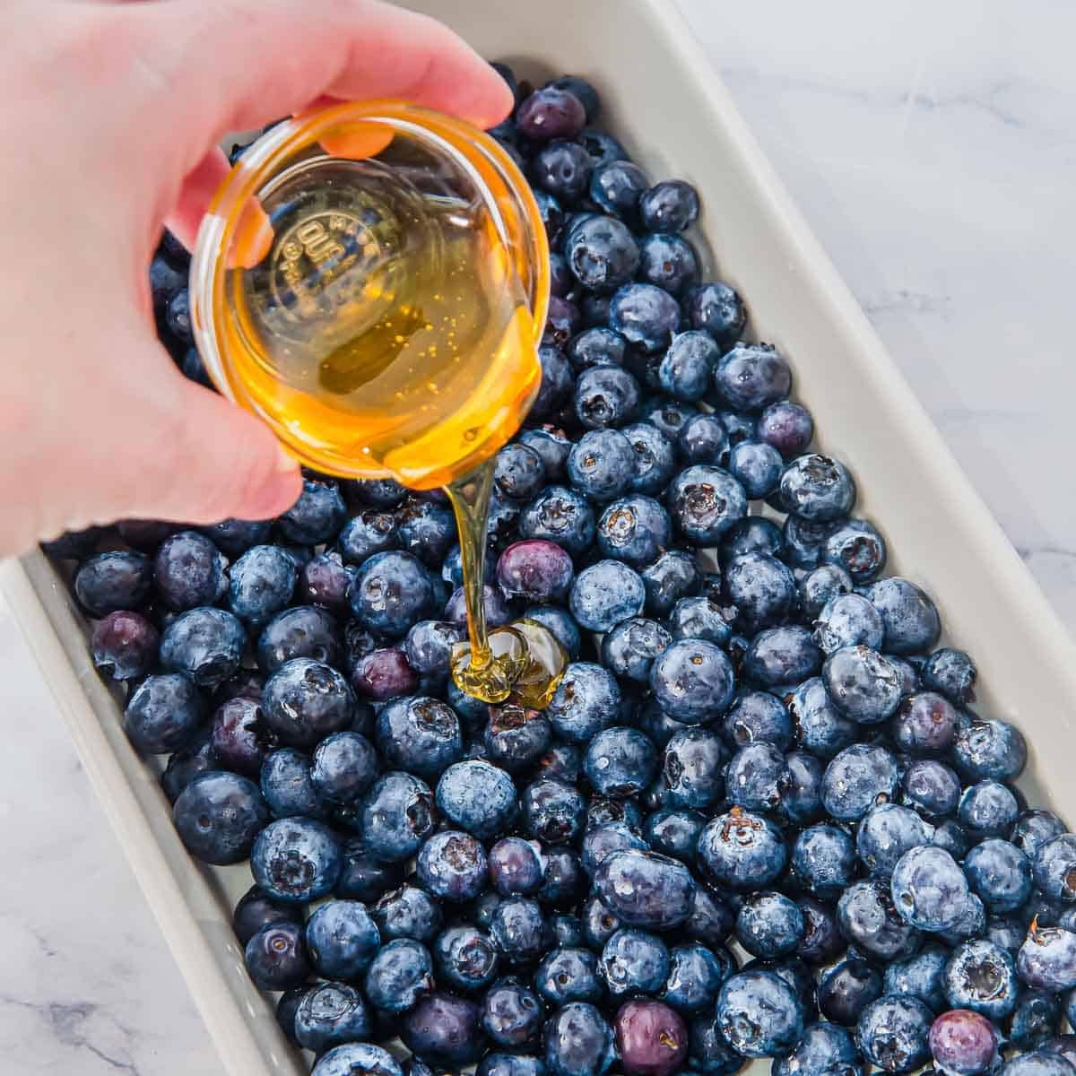 A hand pours golden honey from a small glass bowl over fresh blueberries arranged in a rectangular white baking dish.