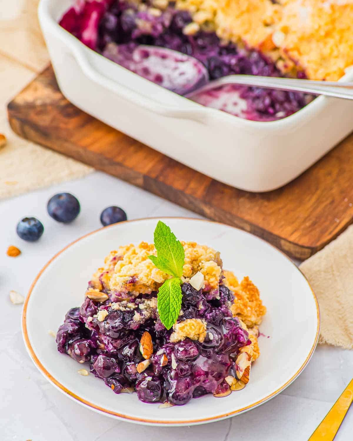 A serving of blueberry cobbler topped with a mint sprig sits on a white plate, with a baking dish of the dessert and a spoon in the background. Blueberries and chopped nuts are scattered around.