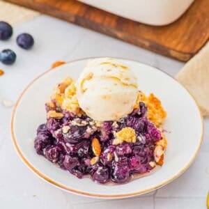 A white plate with a serving of blueberry cobbler topped with a scoop of vanilla ice cream, garnished with sliced almonds. Fresh blueberries and a wooden board are in the background.