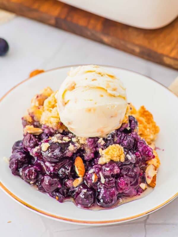 A white plate with a serving of blueberry cobbler topped with a scoop of vanilla ice cream, garnished with sliced almonds. Fresh blueberries and a wooden board are in the background.