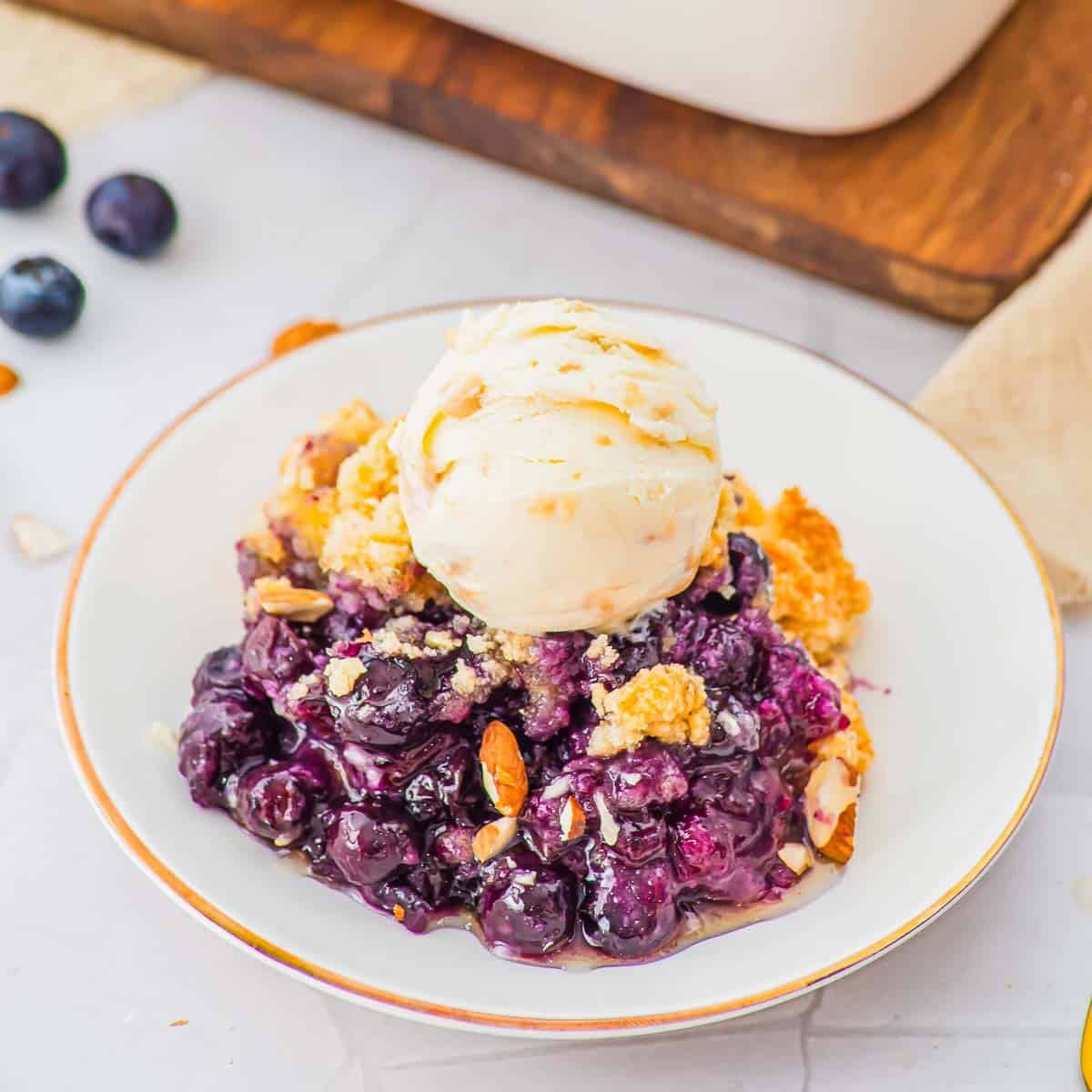 A white plate with a serving of blueberry cobbler topped with a scoop of vanilla ice cream, garnished with sliced almonds. Fresh blueberries and a wooden board are in the background.